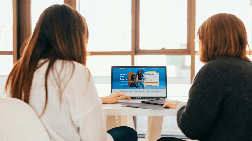 Twee vrouwen zitten aan een tafel en kijken naar een laptop met daarop een Zorgkiezer website. Ze kiezen een zorgverzekering. Ze zitten in een lichte kamer met grote ramen.